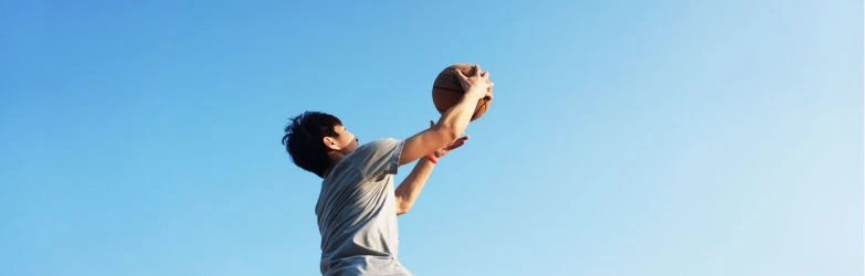Ein Basketballspieler springt vor einem klaren blauen Himmel hoch, um den Ball ins Tor zu werfen